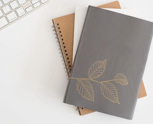 Stack of notebooks with leaf illustration gold foil stamped on cover laying on white computer desk