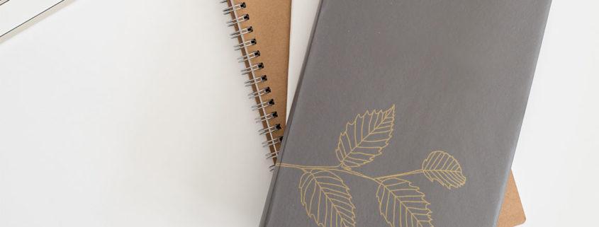 Stack of notebooks with leaf illustration gold foil stamped on cover laying on white computer desk